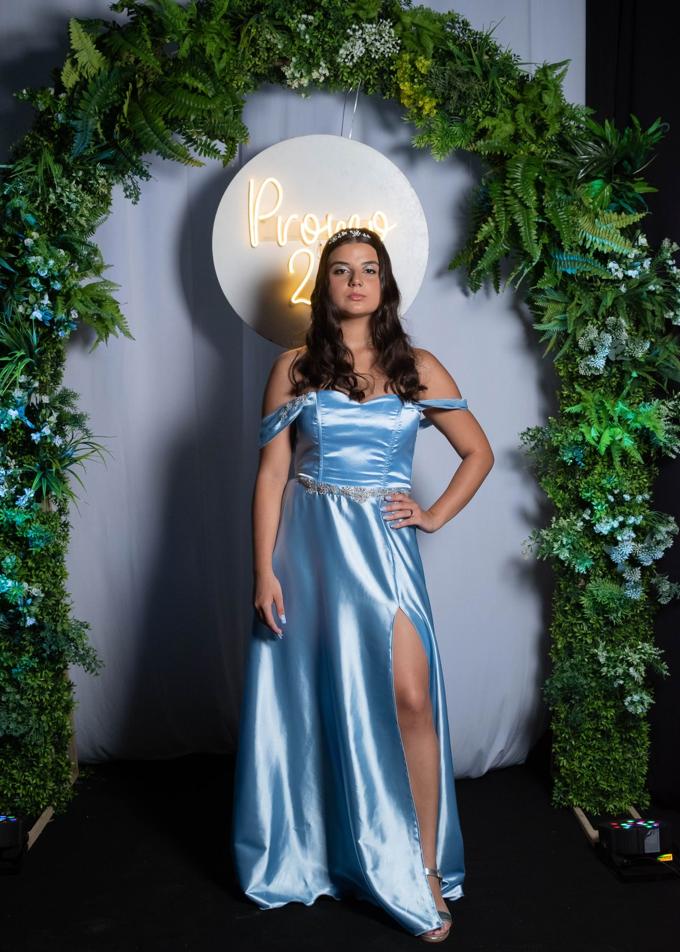 A young woman in a blue satin gown and tiara standing under a floral prom arch