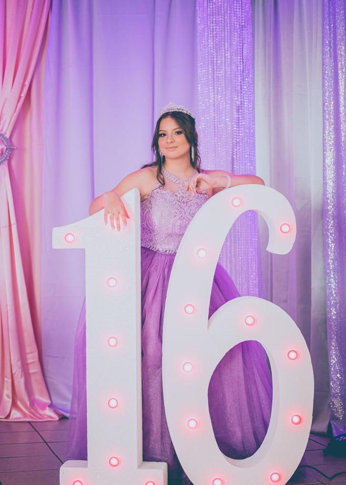 A teenager in a lilac gown and tiara beside a lit marquee sixteen at her Sweet Sixteen party